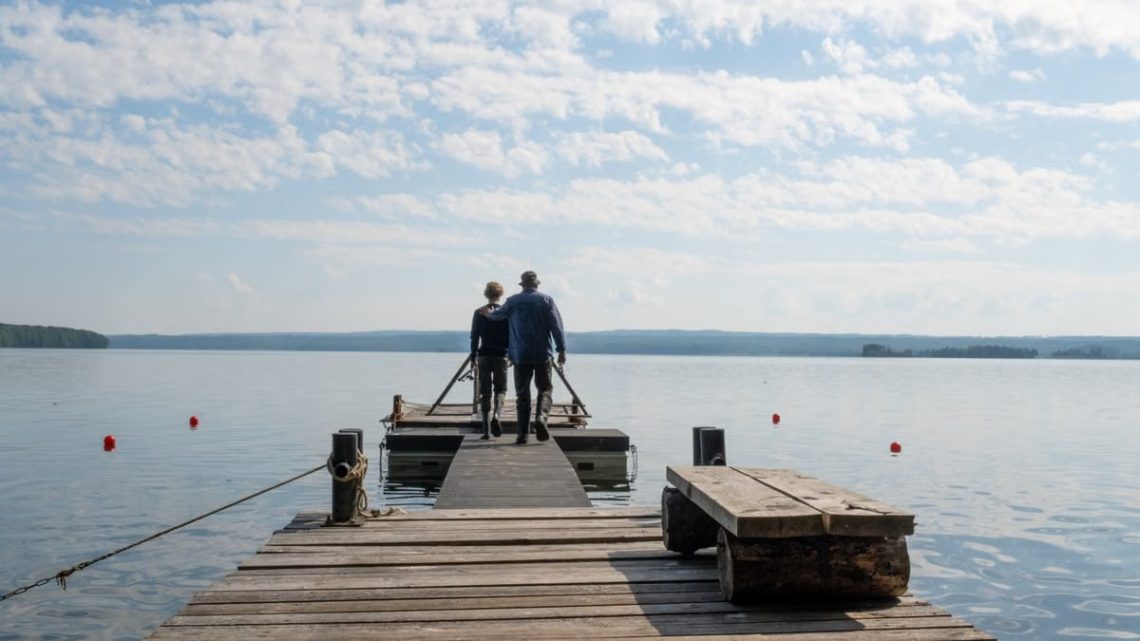 A couple walks on a wooden pier over a serene lake, surrounded by nature and clear skies.