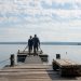 A couple walks on a wooden pier over a serene lake, surrounded by nature and clear skies.